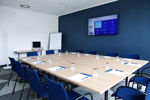 Una sala de conferencias con una mesa larga, sillas azules, una pizarra y una pantalla mostrando una presentación.