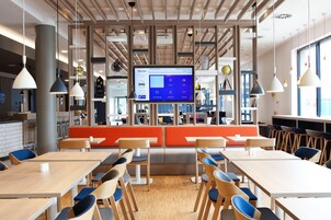 Un comedor moderno con mesas y sillas de madera, una zona de estar anaranjada en el centro y una pantalla digital mostrando un menú.