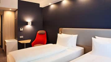 A hotel room with a large bed, a red chair, a small table, and a wall-mounted light.