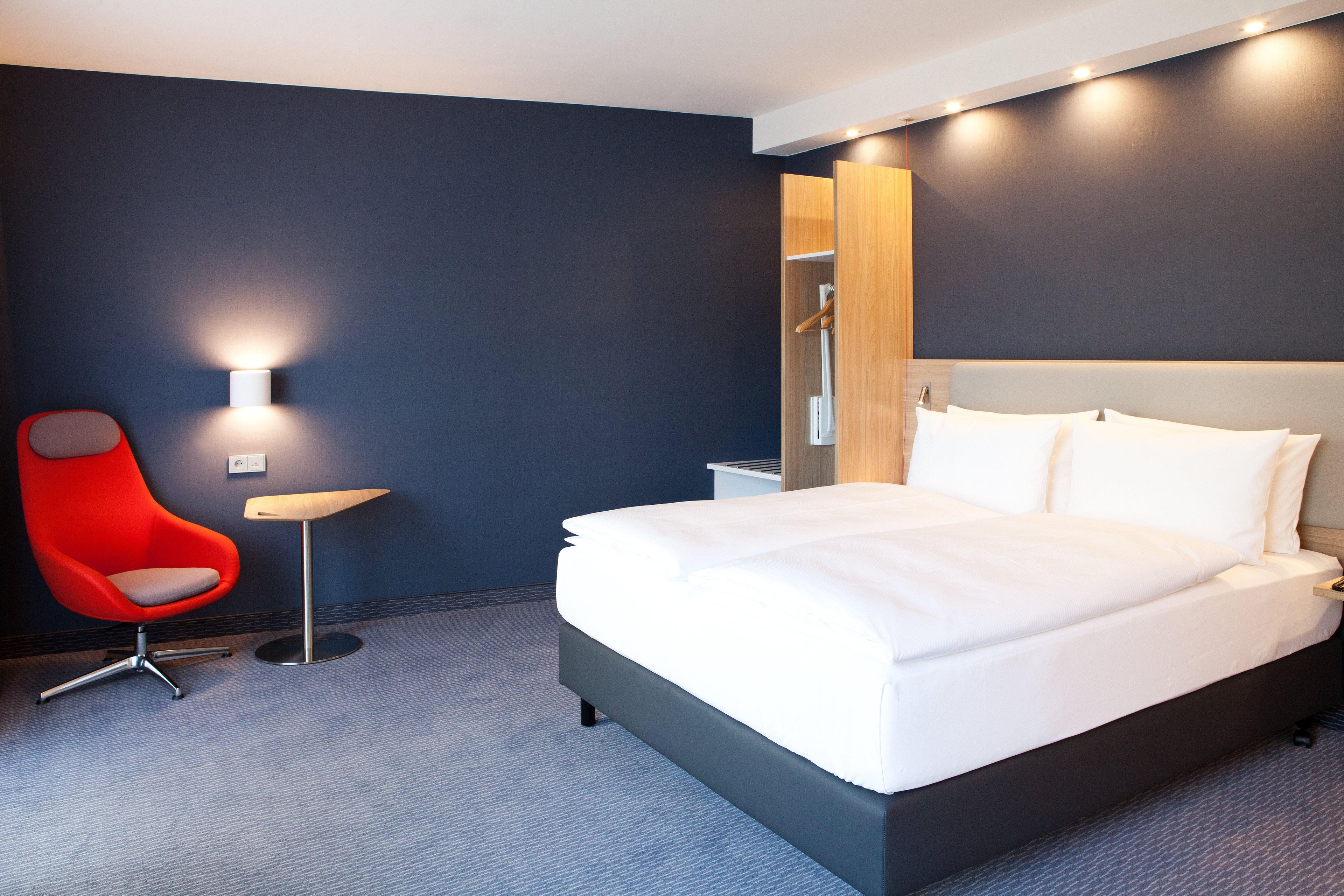Una habitación de hotel moderna con una cama grande, una silla roja, una mesita pequeña y una lámpara fijada a la pared.