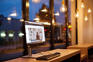Una computadora de escritorio con monitor mostrando una página web, conectada a un teclado y ratón, sobre una mesa de madera cerca de una ventana con bombillas colgantes.