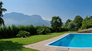 Una piscina rectangular con pared de piedra y escalera metálica en un lado, rodeada de césped cuidado y follaje, con montañas al fondo.