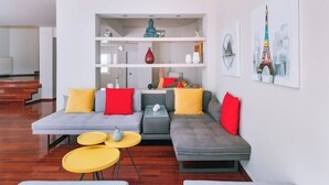 A modern living room with a grey sectional sofa, yellow and red cushions, a white shelving unit with decorative items, and a painting of the Eiffel Tower. - Villa Politia   Luxury Villa with a Spectacular City View (Athens)
