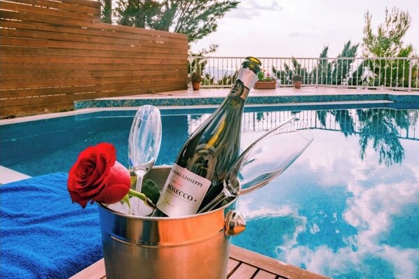 A metal bucket with a bottle of champagne and a glass, placed on a blue towel by a pool.