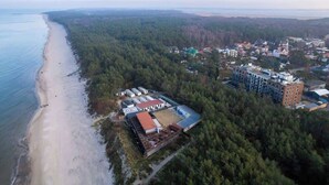 An aerial view of a coastal area with a beach, dense forest, and a cluster of buildings including a large multi-story structure. - Bright apartment with balcony | Right on the beach in Miedzywodzie (Międzywodzie)