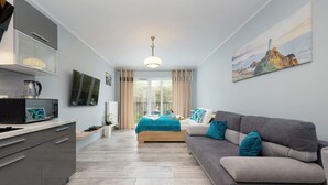 A modern hotel room with a grey sofa, a bed with a blue bedspread, a flat-screen TV, a coffee maker, and a painting of a lighthouse on the wall. - Bright apartment with balcony | Right on the beach in Miedzywodzie (Międzywodzie)