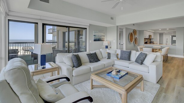 Ett rymligt vardagsrum med en stor vit soffa, ett soffbord i glas och utsikt över havet genom stora skjutdörrar.