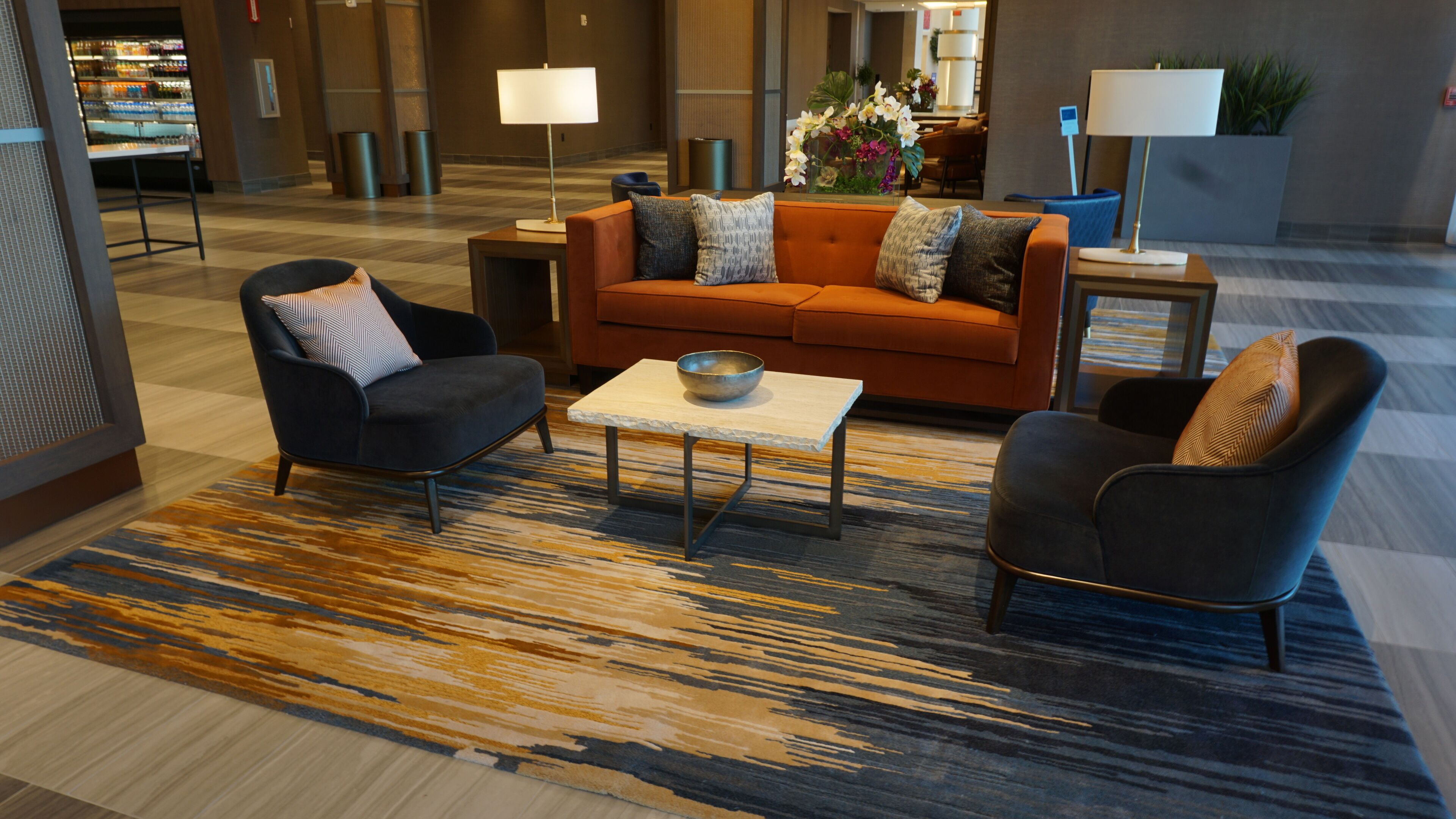 Um lobby moderno com um sofá, poltronas e uma mesa de centro. Há uma geladeira e um espaço de refeições ao fundo.
