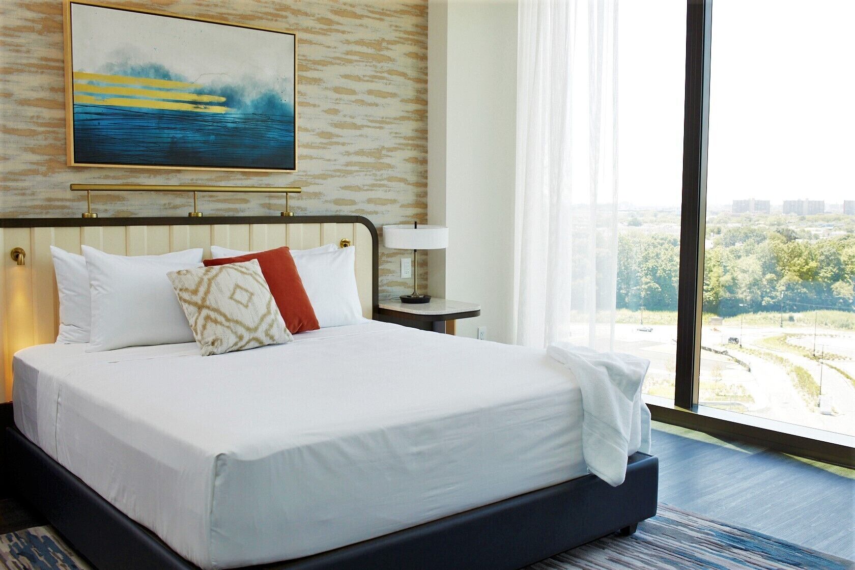 A hotel room with a large bed, a painting above the bed, a nightstand with a lamp, and a view of a cityscape through large windows.