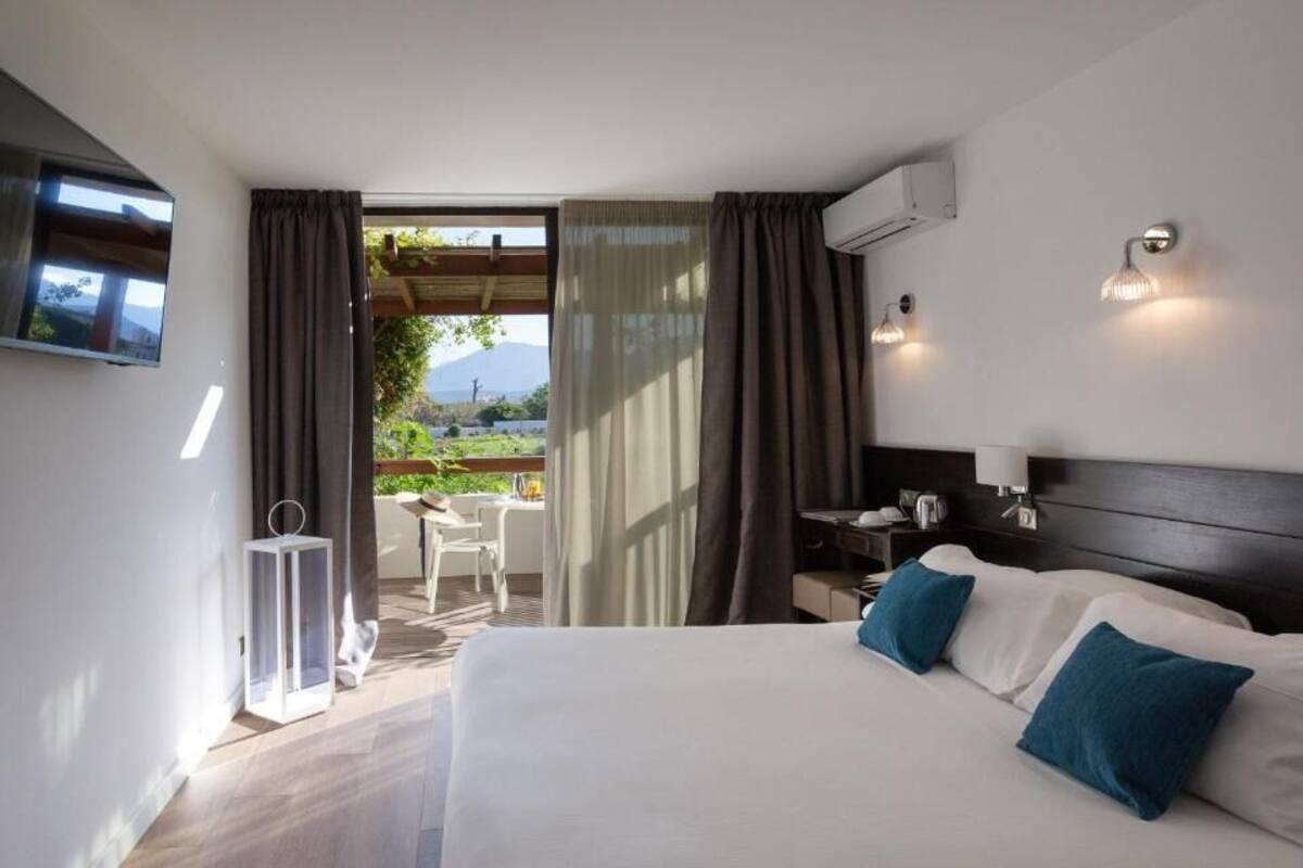 A hotel room with a bed, a TV mounted on the wall, a small table with chairs, and a view of the outdoors.