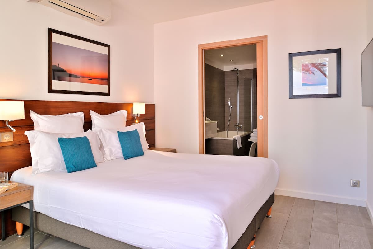 A hotel room with a large bed, a wooden headboard, a nightstand, a framed picture of a sunset, a bathroom with a shower, and a framed picture on the wall.