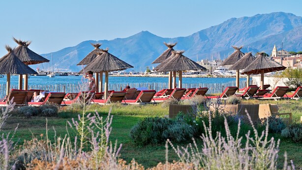 A beachside area with rows of sun loungers and thatched umbrellas, a clear view of the sea, and mountains in the background. - La Roya (Saint-Florent)