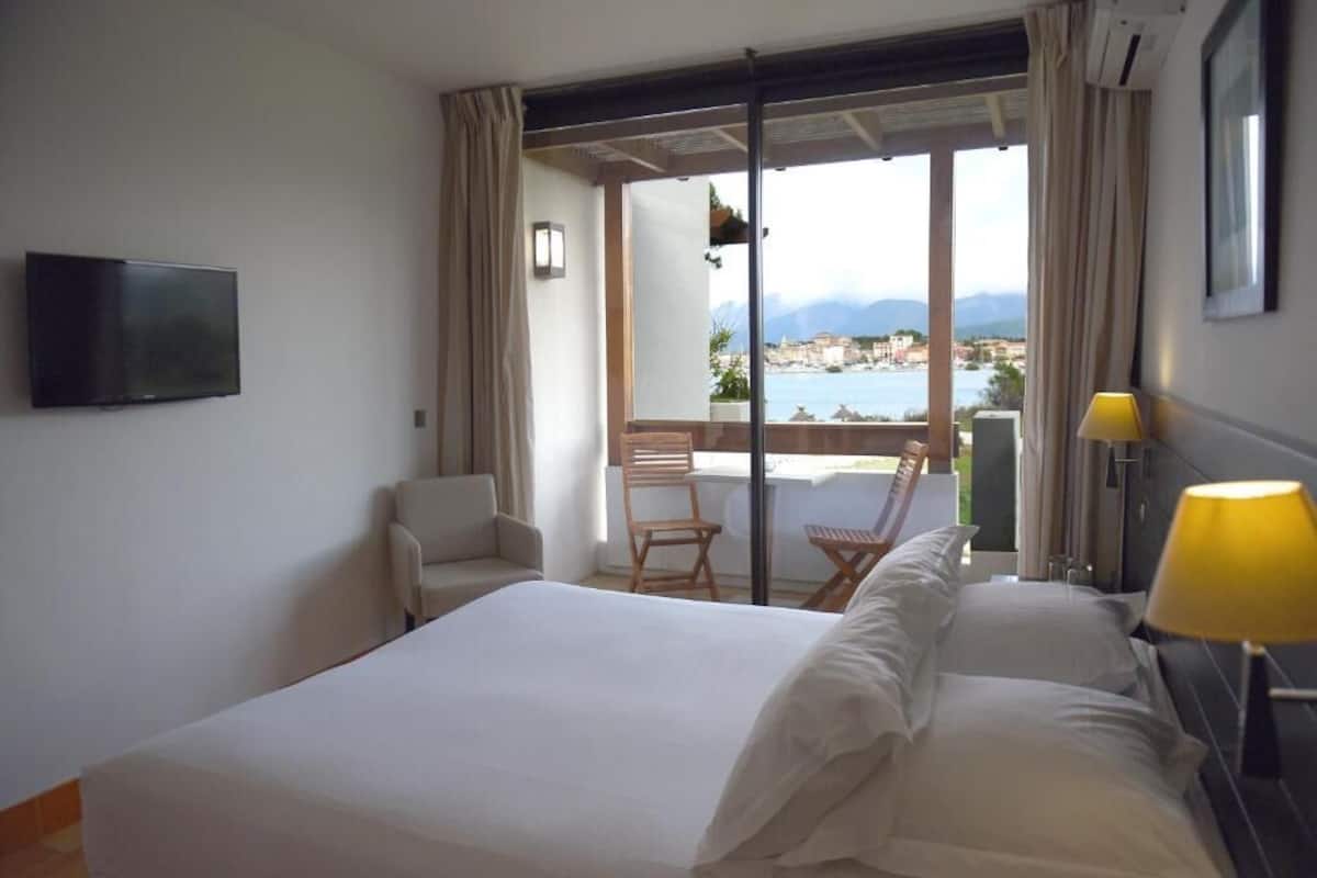 A hotel room with a bed, a TV, a chair, a table, and a view of the sea.