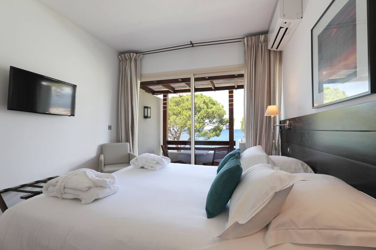 A hotel room with a bed, a TV, a window with curtains, and a view of the outdoors.
