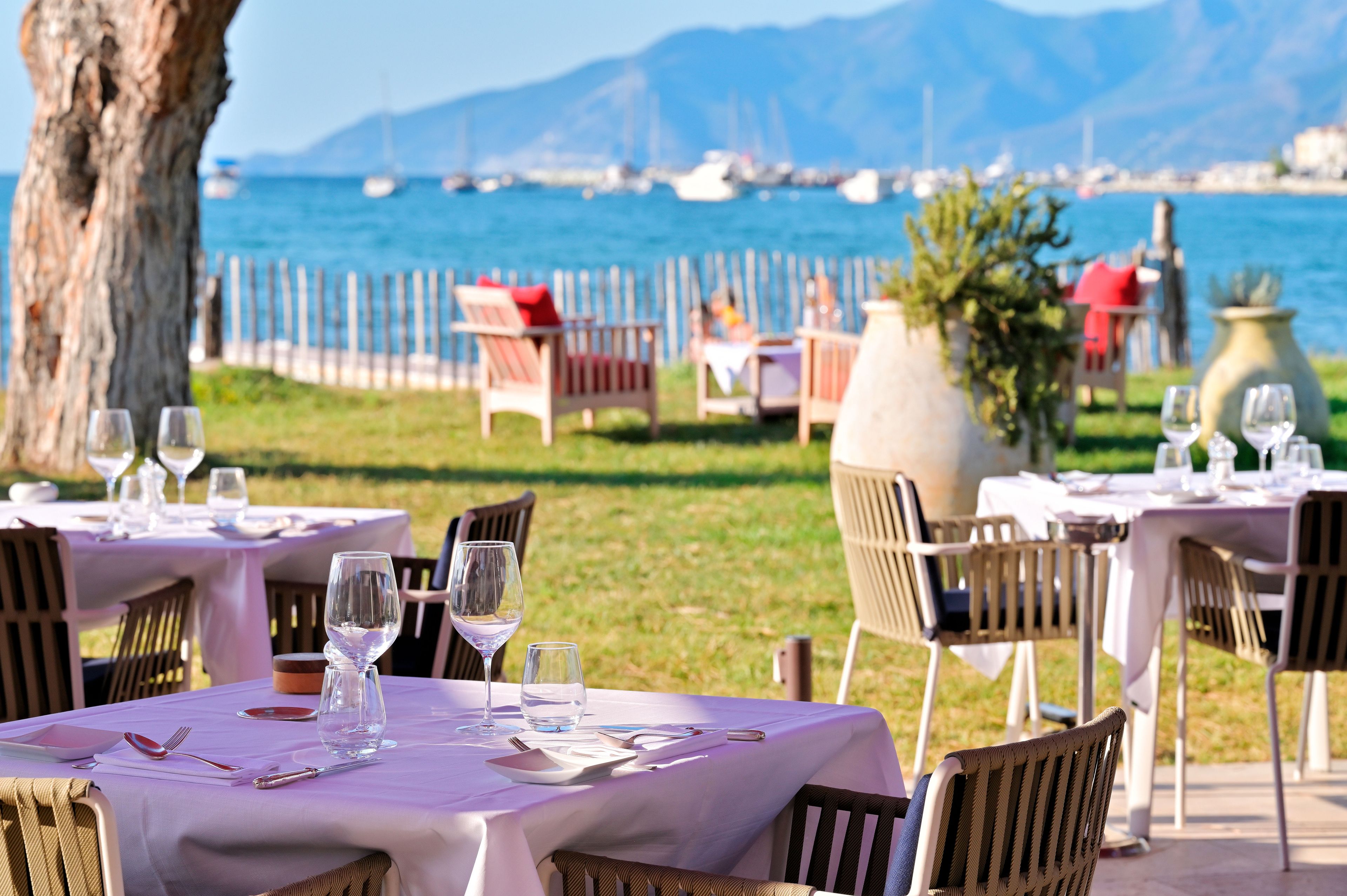 Outdoor dining tables with chairs, a view of the sea, and a mountainous backdrop.