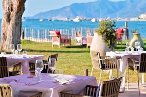 Mesas y sillas para comer al aire libre, con vista al mar y montañas al fondo.