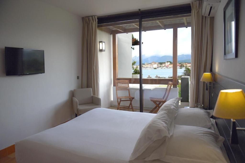 A hotel room with a bed, a TV, a chair, a table, and a view of the sea.