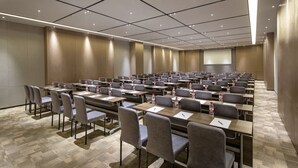 Uma sala de conferências com várias mesas e cadeiras, equipada com garrafas de água e canetas, e uma tela de projeção na parede.