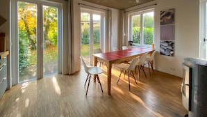 Un vaste espace dédié à la salle à manger, avec une table en bois et des chaises blanches, de grandes portes-fenêtres coulissantes donnant sur un jardin, et une cuisine moderne équipée d’une cuisinière.