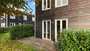 Un immeuble résidentiel moderne à deux étages, doté d’une façade en bois foncé, de fenêtres à cadre blanc et d’un jardin bien entretenu aux feuilles jaunes.