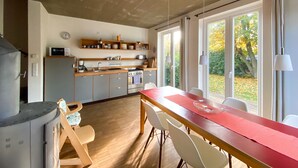 Une cuisine moderne avec des meubles en bois, une table à manger avec des chaises et de grandes fenêtres donnant sur l’extérieur.