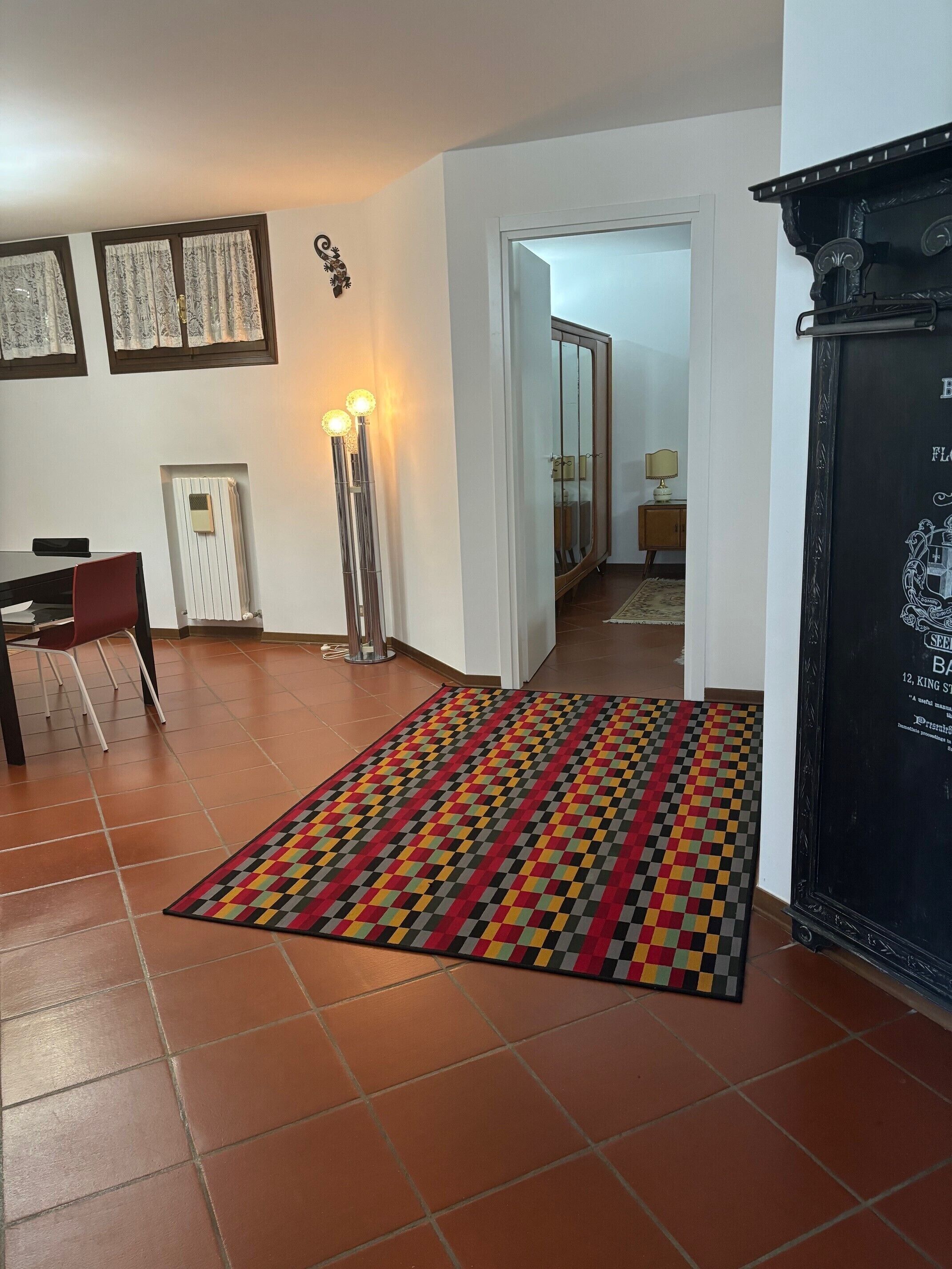 Ein Zimmer mit Fliesenboden, einem gemusterten Teppich, einem Schreibtisch mit Stuhl, einem Heizkörper und einer hohen Stehlampe.