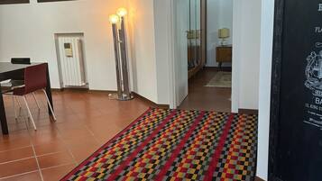 Ein Zimmer mit Fliesenboden, einem gemusterten Teppich, einem Schreibtisch mit Stuhl, einem Heizkörper und einer hohen Stehlampe.