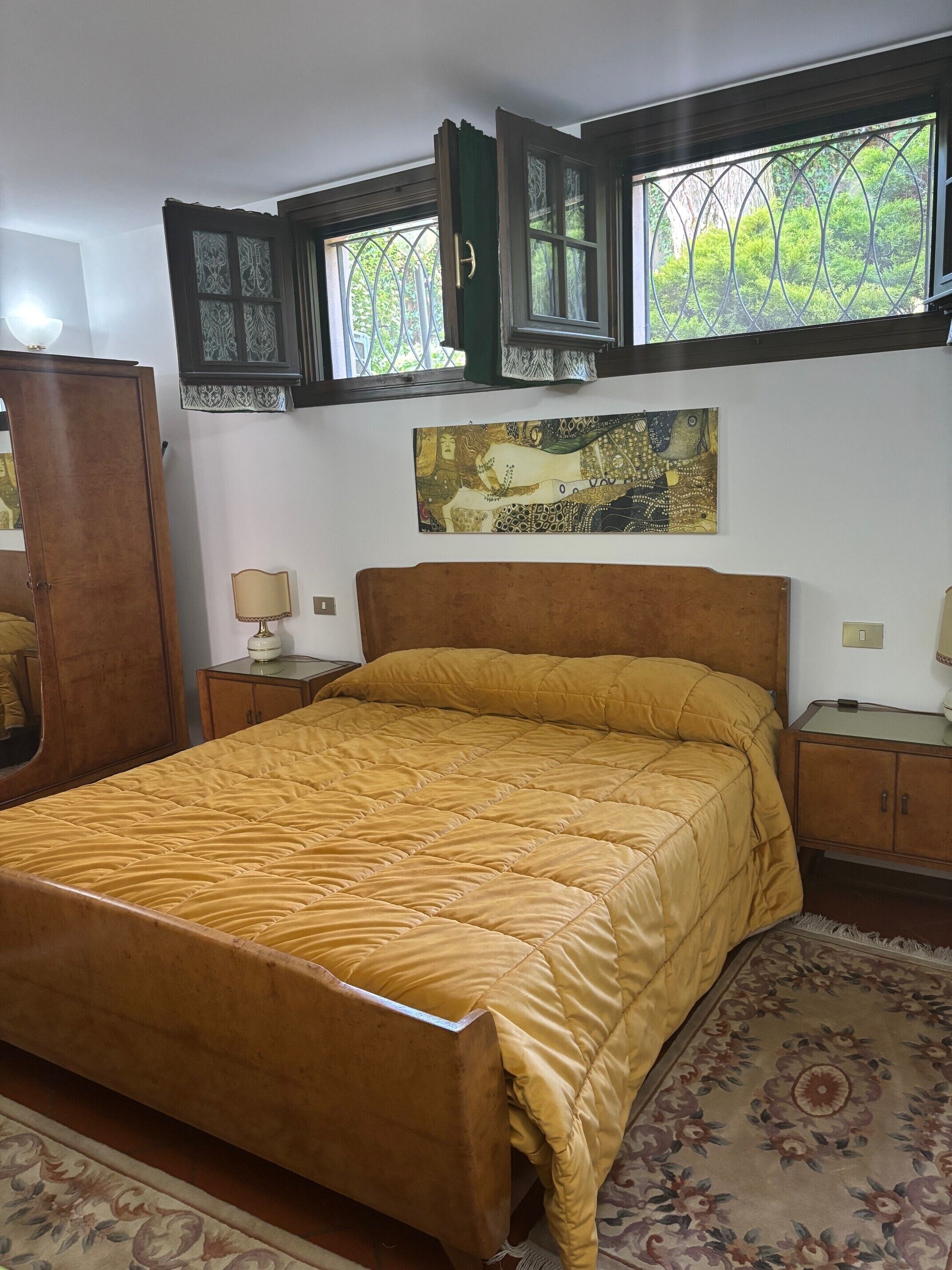 A bedroom with a bed, wooden headboard, two nightstands, a mirror, a window with curtains, and a painting on the wall.