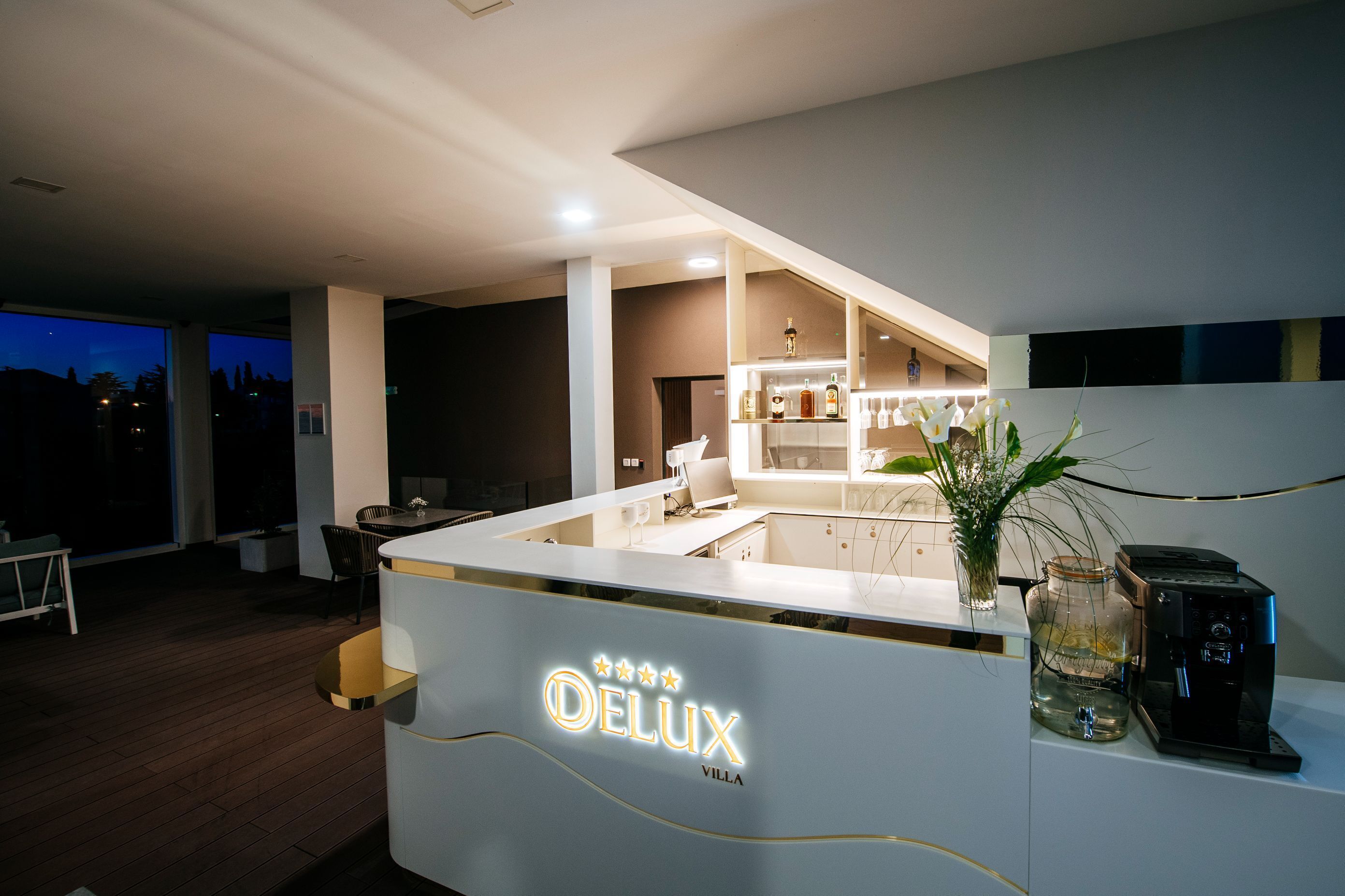 Ein moderner Empfangsbereich mit einer Theke mit der Aufschrift "DELUXE VILLA", einer Kaffeemaschine und einer Vase mit Blumen.