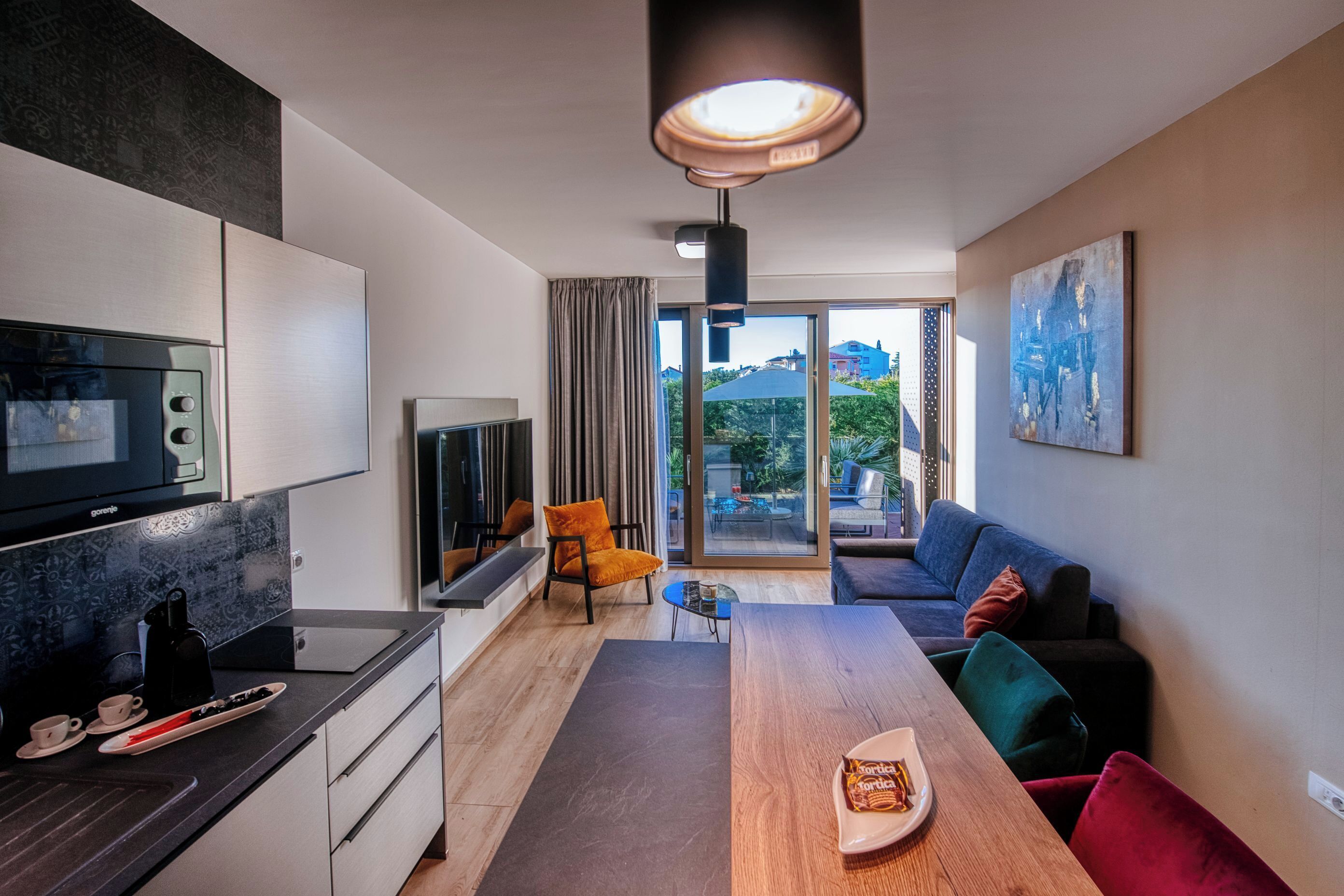 Uma sala moderna com uma mesa de jantar de madeira, um sofá, uma TV e uma área de cozinha com micro-ondas e máquina de café.
