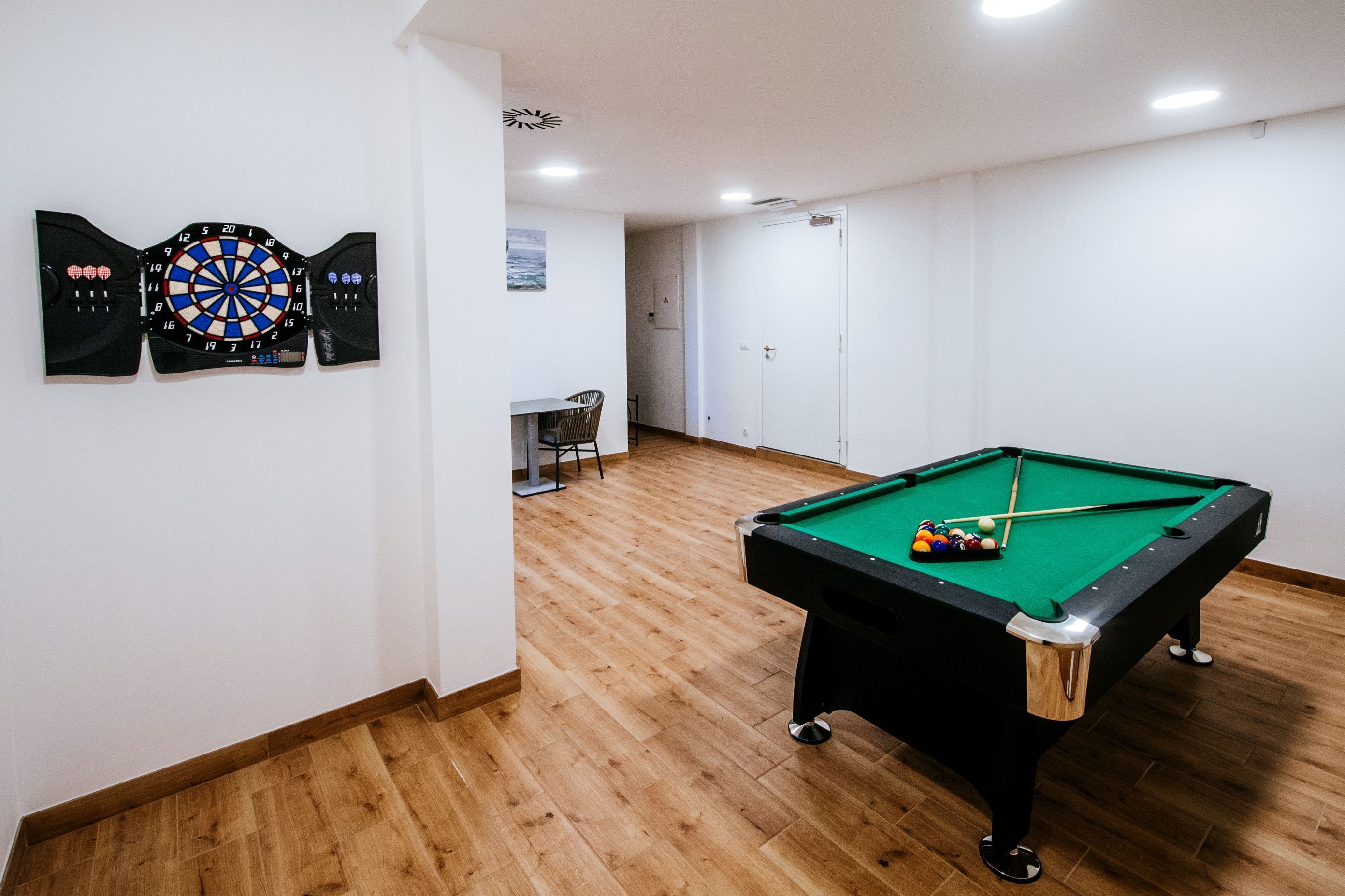 Ein Billardtisch mit Kugeln, eine Dartscheibe und ein Stuhl in einem Raum mit Holzboden und weißen Wänden.