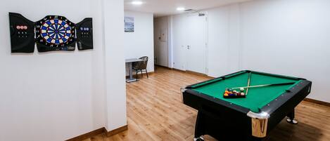 Ein Billardtisch mit Kugeln, eine Dartscheibe und ein Stuhl in einem Raum mit Holzboden und weißen Wänden.