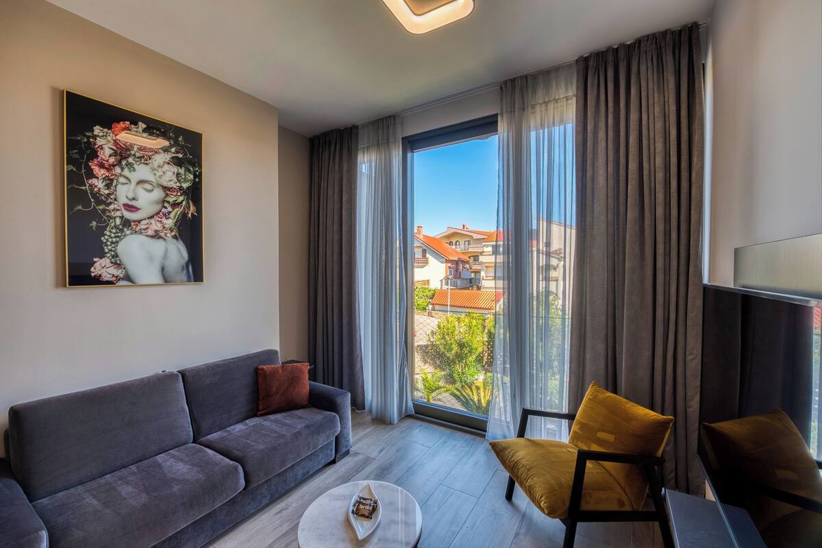 A modern living room with a grey sofa, a round coffee table, and a yellow armchair. A large window with curtains offers a view of buildings and greenery outside.