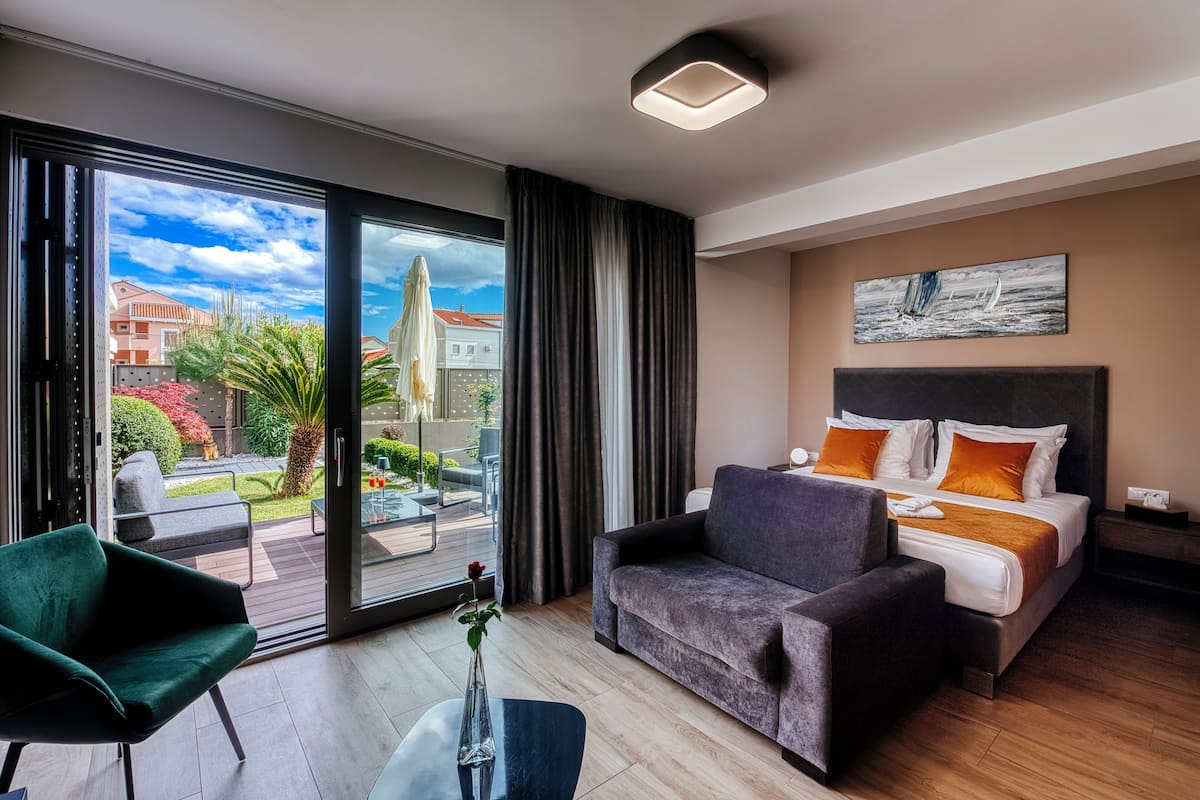 A modern hotel room with a large sliding glass door leading to a balcony, a comfortable bed with orange pillows, a dark green armchair, and a small table with a vase.
