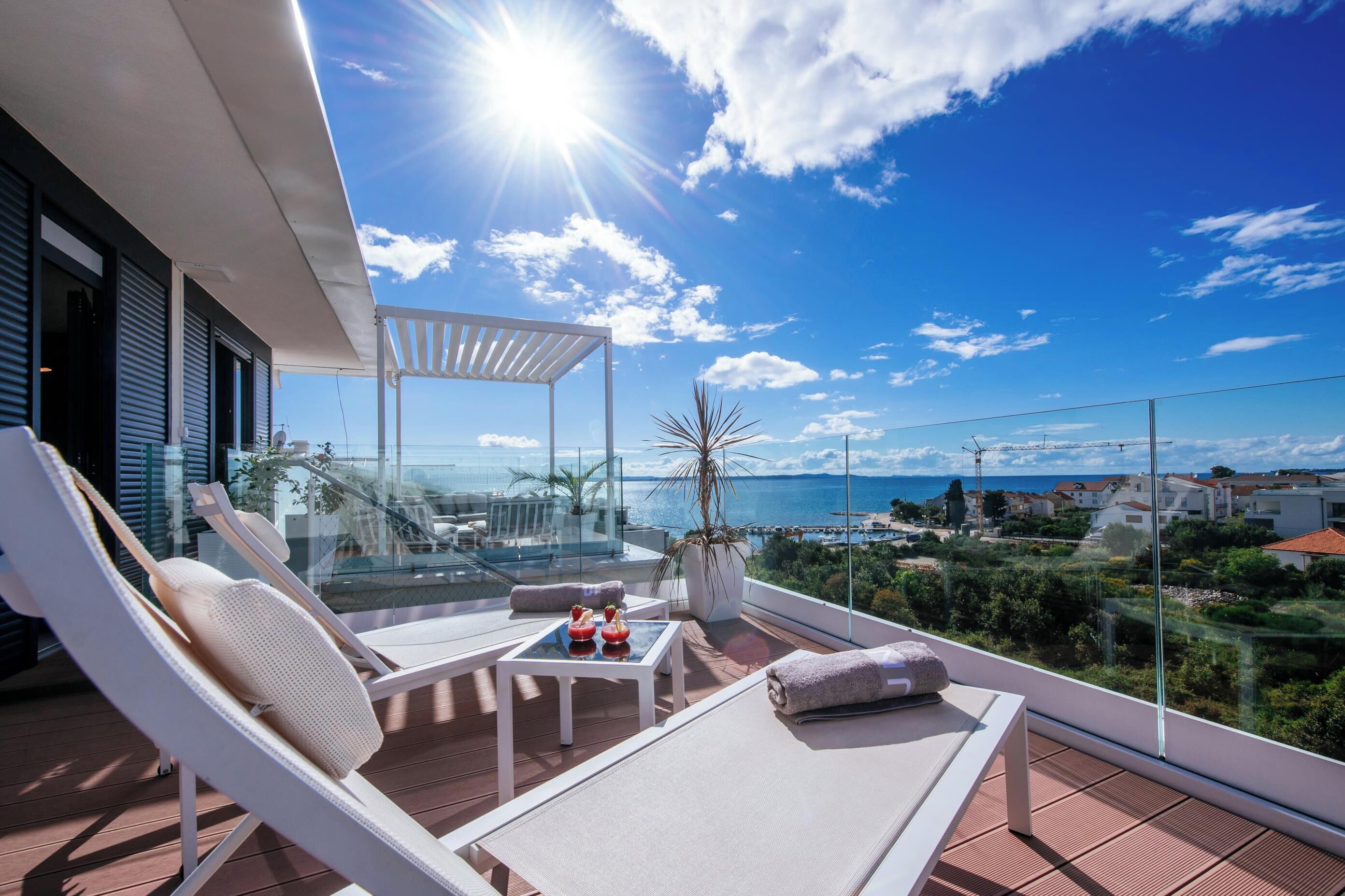 Un balcon ensoleillé avec des fauteuils de jardin blancs, une petite table agrémentée d’objets décoratifs, et une vue sur la mer et les bâtiments environnants.