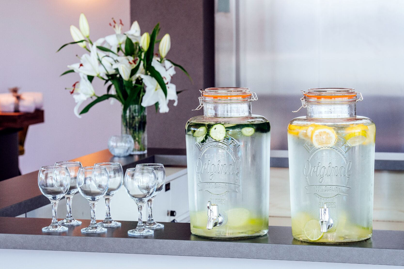 Zwei Glasflaschen mit Ausguss, gefüllt mit einer klaren Flüssigkeit und mit Zitronenscheiben und Gurkenscheiben verziert, stehen auf einer Theke, auf der leere Weingläser Platz finden.