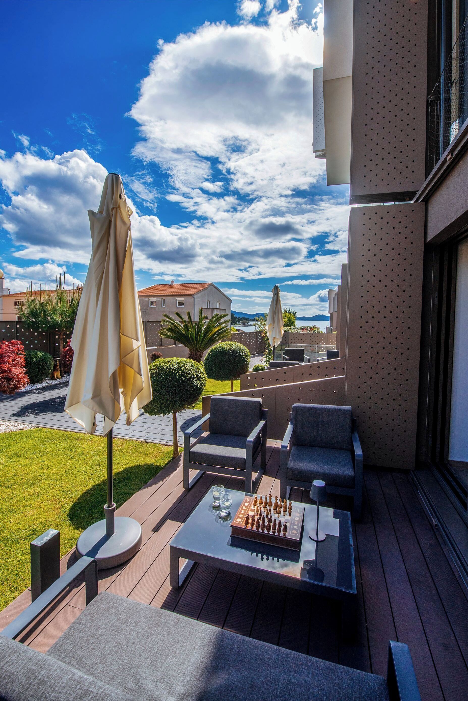A deck with outdoor furniture, including chairs and a table with a chess set, overlooking a garden and buildings.