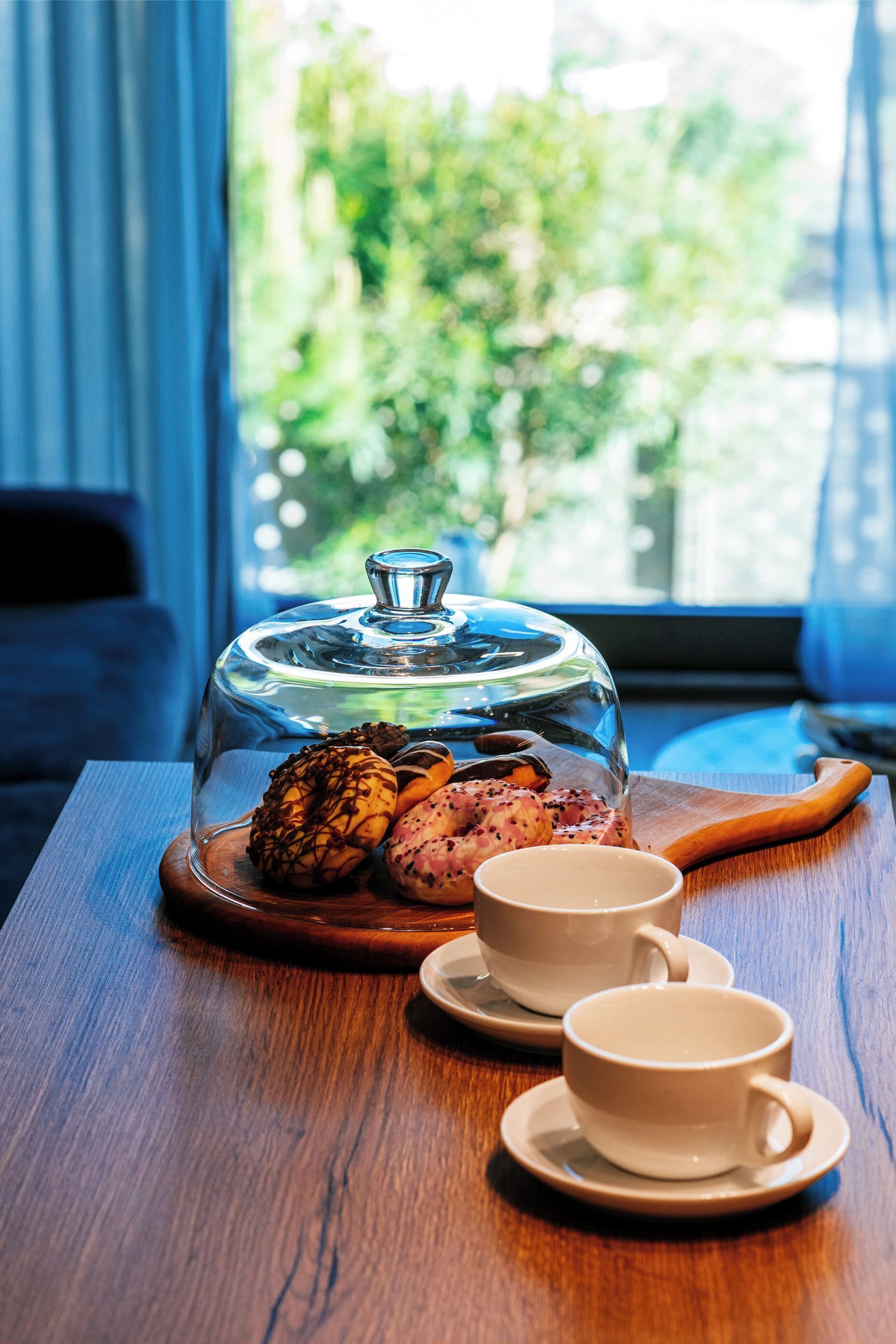 A wooden table with a glass dome-covered dessert, two white cups, and a wooden spoon.