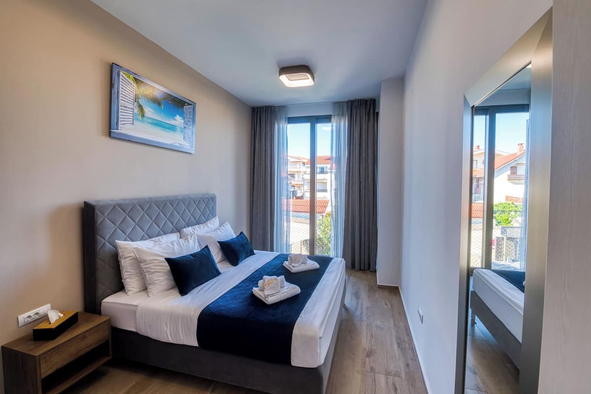 A modern hotel room with a large bed, a bedside table, a painting on the wall, and a view of the outside.