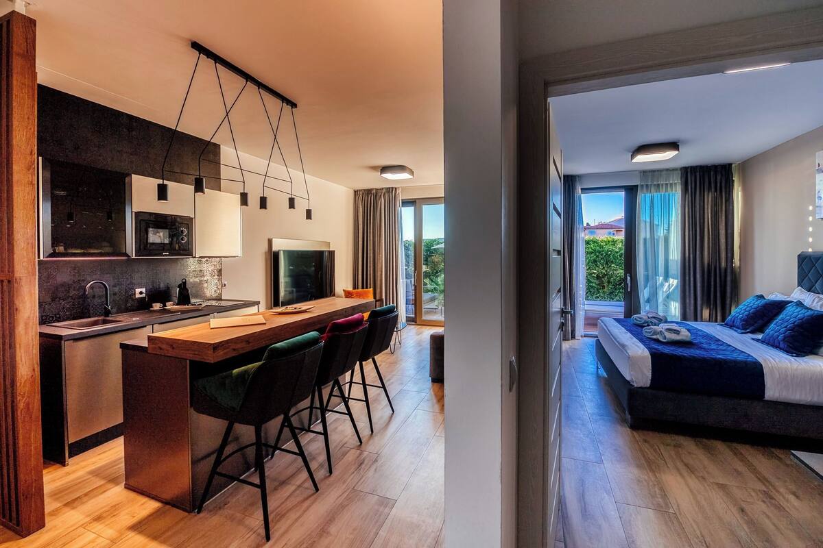 A modern hotel room with a kitchen area, a dining space, and a bedroom with a balcony.