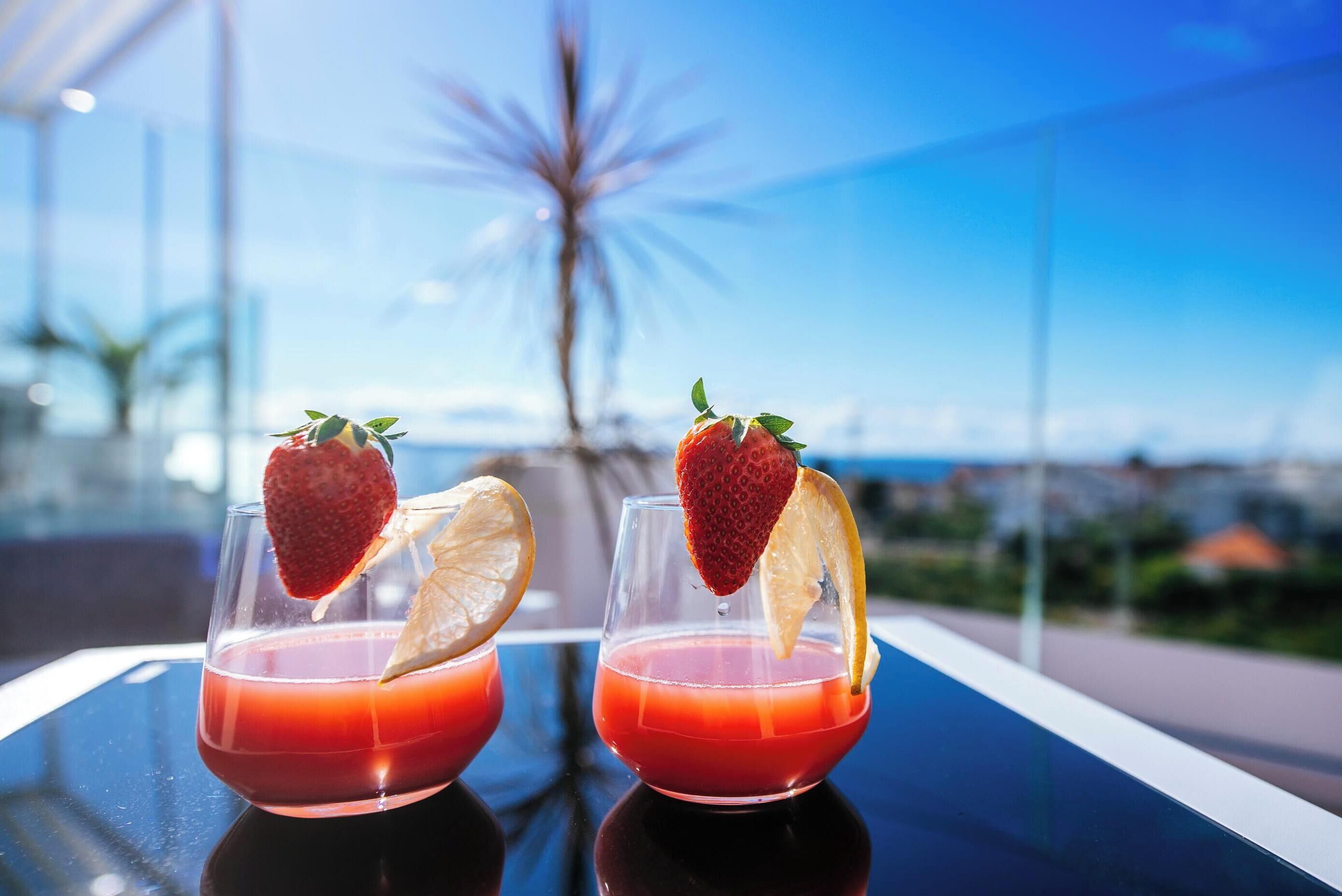 Two glasses of red drink with strawberries and lemon slices on a reflective surface.
