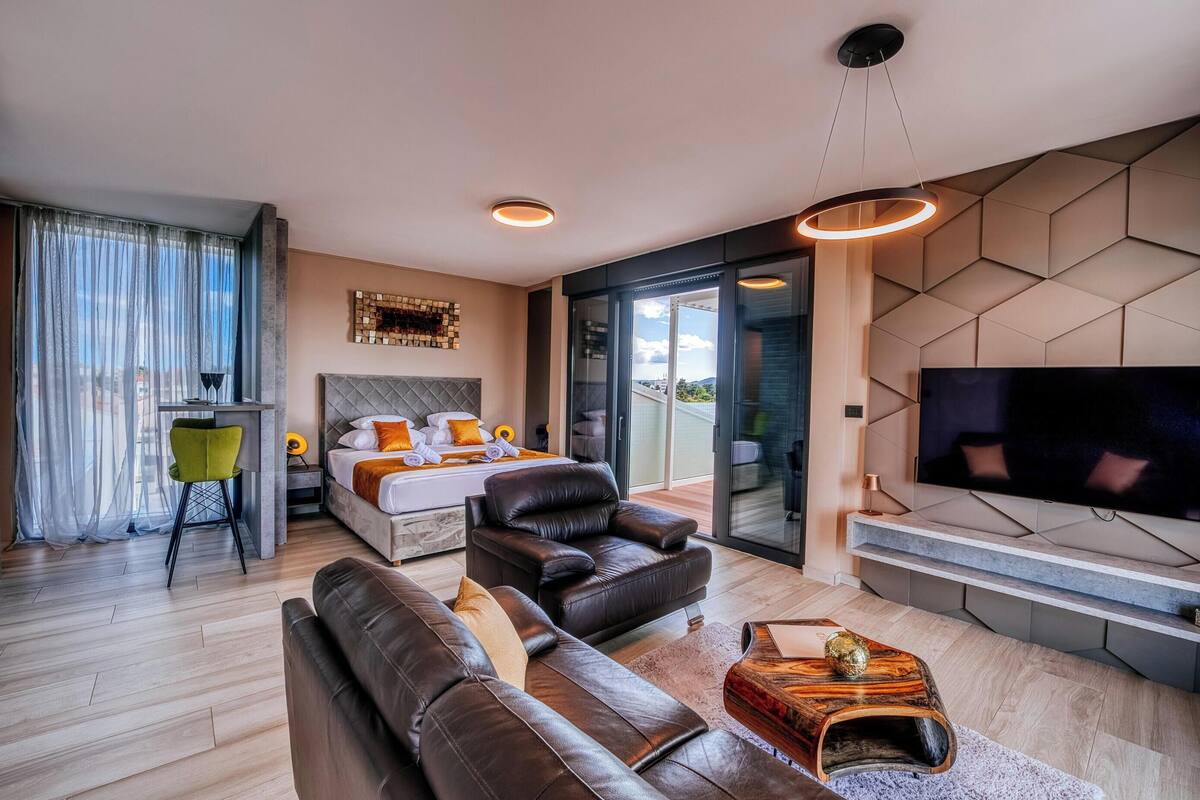 A modern hotel room with a bed, leather sofa, a TV, and a dining area.