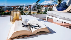 An open book, two glasses of white wine, a pair of glasses, and a straw hat on a white table with a view of a cityscape.
