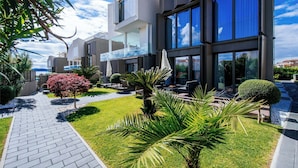 A modern multi-story building with a paved walkway, well-maintained lawn, and various plants.