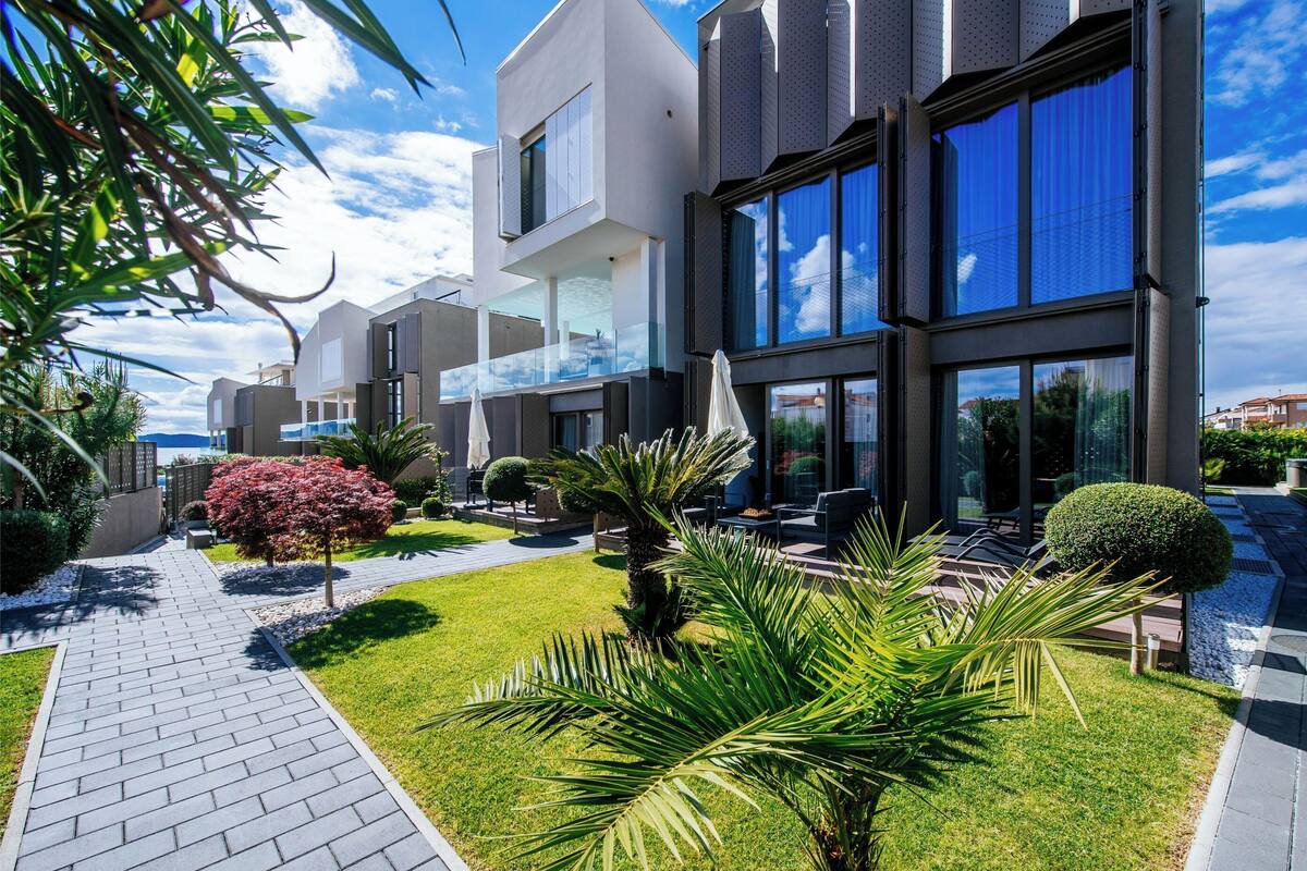 A modern multi-story building with a paved walkway, well-maintained lawn, and various plants.