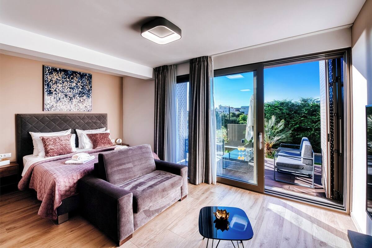 A modern hotel room with a bed, a sofa, a small table, and a sliding glass door leading to an outdoor area.