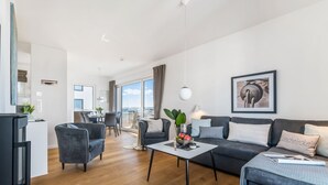 A modern living room with a sofa, coffee table, and dining area. There is a large window providing a view of the city. - Penthouse Sonnendeck 22 - Balcony, Barrel Sauna, Grill,Fireplace,Washing Machine (Olpenitz)