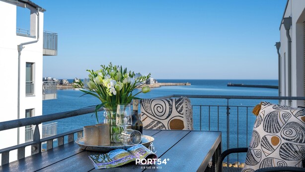 A balcony with a table set for dining, a vase with flowers, and a view of the sea. - Penthouse Sonnendeck 22 - Balcony, Barrel Sauna, Grill,Fireplace,Washing Machine (Olpenitz)