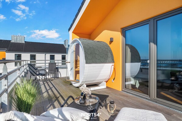 A modern outdoor deck with a unique cylindrical structure, a lounge area with a tray table, and a glass-enclosed room. - Penthouse Sonnendeck 22 - Balcony, Barrel Sauna, Grill,Fireplace,Washing Machine (Olpenitz)