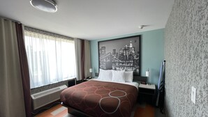 Ein Hotelzimmer mit Bett, einem Stadtbildgemälde, einem Heizkörper, einer Uhr und einem Fenster mit Vorhängen.